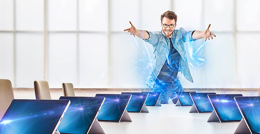 Lächelnder Mann mit ausgebreiteten Armen steht vor einer Reihe von Laptops in einem Konferenzraum