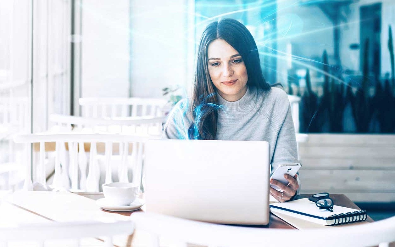 Frau sitzt in einem Café und arbeitet an ihrem Laptop, während sie ihr Smartphone in der Hand hält