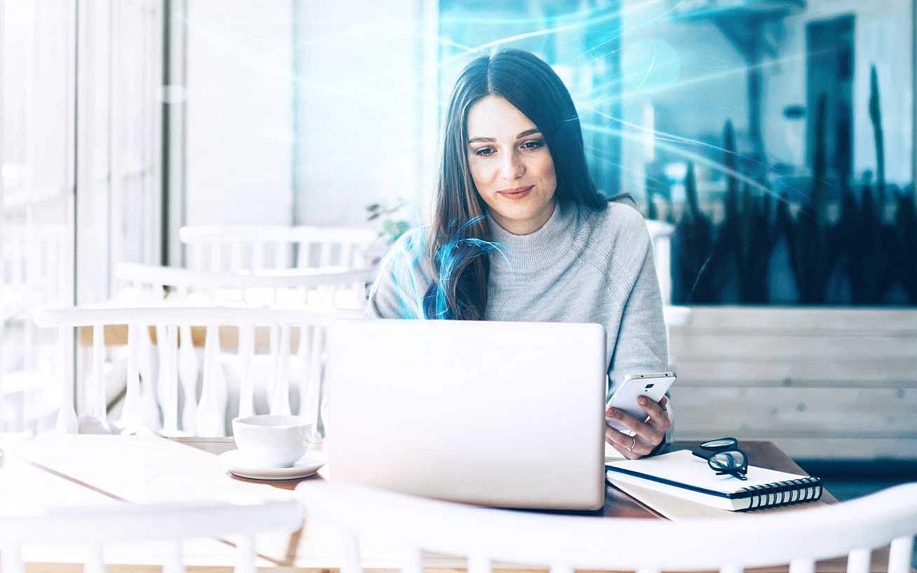 Frau sitzt in einem Café und arbeitet an ihrem Laptop, während sie ihr Smartphone in der Hand hält