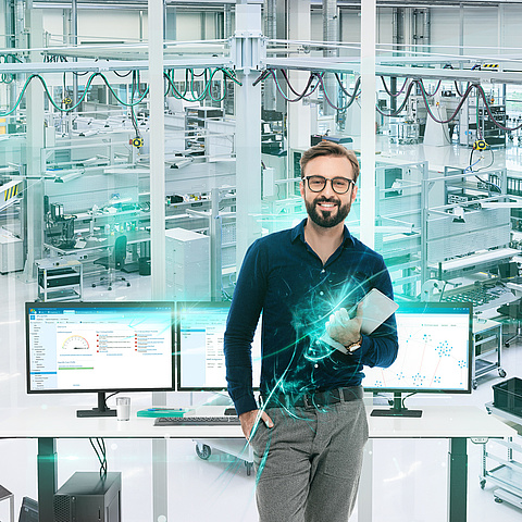 Ein Mann steht in einem modernen industriellen Umfeld mit einem Tablet in der Hand und zwei Bildschirmen vor sich, die die baramundi Management Suite zeigen